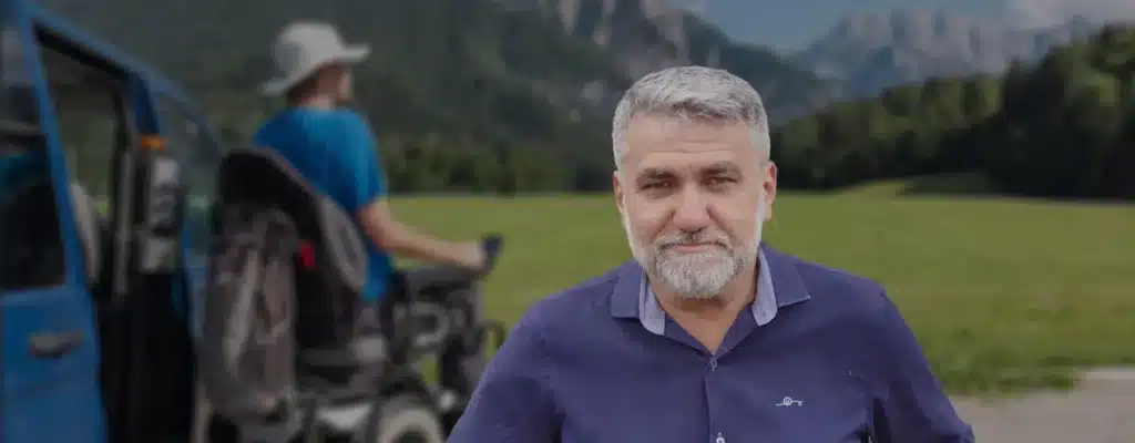 Paisagem com uma montanha ao fundo, um campo verde, um cadeirante saindo de seu carro acessivel, a frente imagem de Marcello Patelli, homem branco, cabelos grisalhos, usando uma camisa polo azul.